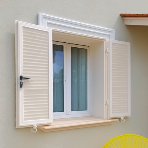 Finestra blindata a due ante con infissi in acciaio bianco e persiane orientabili, montata su facciata esterna chiara con arredo da esterno.